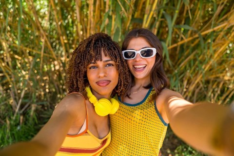 Two smiling girls taking a selfie in front of bamboo plants, wearing yellow clothes and headphones