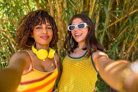 Two cheerful young women with colorful clothes and headphones taking a selfie in front of a bamboo