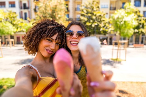 Two happy female friends taking a selfie while showing ice cream cones on a sunny summer day in a
