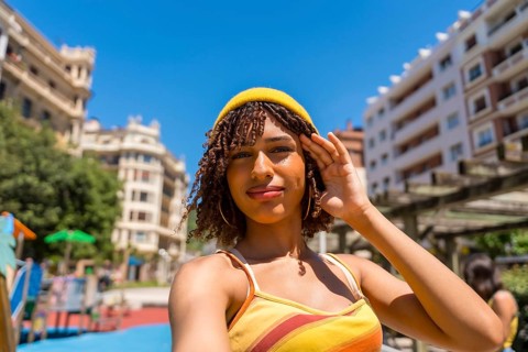 Stylish young woman with curly hair and yellow beanie taking a selfie in a sunny city park,