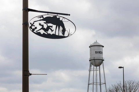 Bandera water tower behind artistic metal sign showing cowboy life on pole in Bandera, Texas, USA