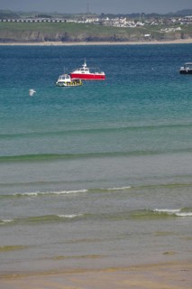 A red boat floats on calm blue water near a wide sandy beach, St Ives, Cornwall, England, United