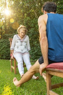 Senior woman exercising her legs with the help of a personal trainer, sitting on chairs in a