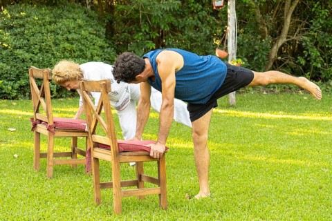 Senior man and woman practicing yoga in garden, doing balancing pose using chairs for support,