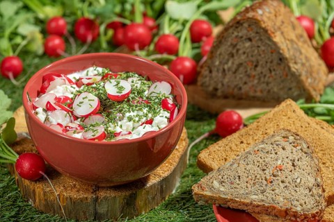 Bread next to a bowl of cream cheese, radishes and herbs on a green meadow