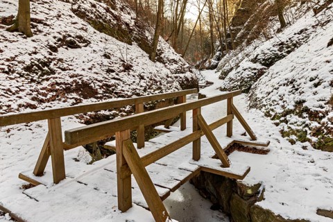 Narrow wooden bridge over a frozen stream in a snowy forest gorge at sunset, The Dragon Gorge near