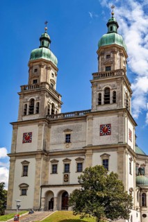 Baroque church with two towers and clocks against a blue sky, surrounded by trees, The town of