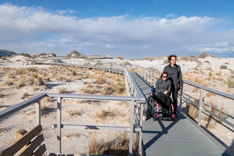 Man and handicapped woman in wheelchair stroll down the Interdune Boardwalk with their ped dog at