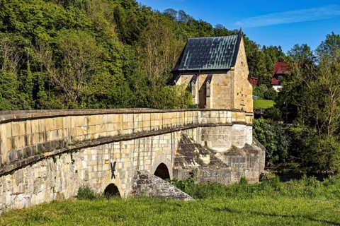 Side view of a historic stone bridge with a chapel and lush vegetation in the background, The