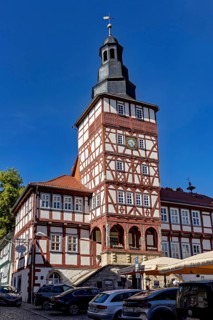 Half-timbered tower with clock surrounded by historic buildings and parked cars on a clear day, The