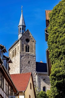 Church with high tower in front of blue sky and surrounding buildings, surrounded by trees, The