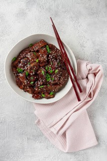 Crispy beef, with green onions and sesame seeds, in a bowl with chopsticks, on a textured surface,