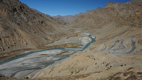 Shimmering blue Indus meanders through a barren valley with serpentines of a road leading down to