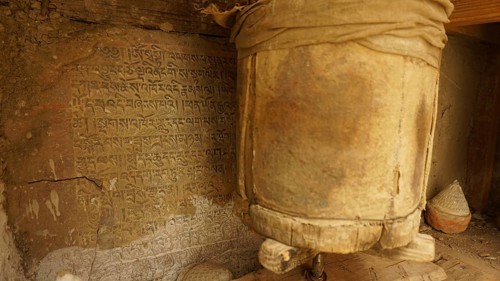 An ancient prayer wheel next to a wall full of inscriptions in a Buddhist context, trekking in