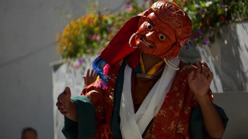 A masked dancer in shades of orange and red with flowers in the background in Leh, trekking in