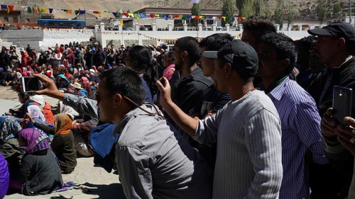 A crowd films an outdoor festival with smartphones, colorful details in the background, Leh,
