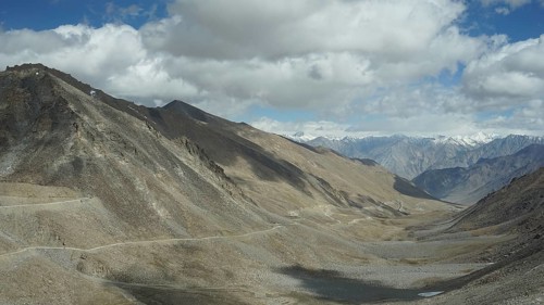 View of a rocky valley with winding mountain road and mountains in the background under slightly