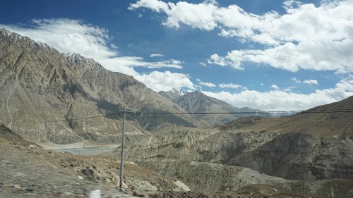 Wide mountain landscape with river valley and power lines under partly cloudy sky, trekking in