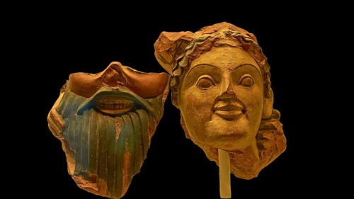 Two ancient clay masks, one with beard and smile, the other with flowing hair, on a dark