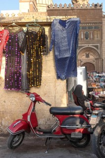 A cool looking red scooter parked amongst a busy street market selling colourful women's dresses