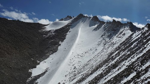 Snowy mountain peak at Khardung-la Pass under clear blue sky with some clouds and rocky landscape,