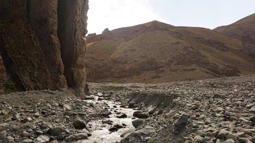 A stream flows through a dry, rocky landscape with high rock walls, trekking at Stok La Pass in