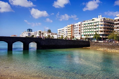 Historic bridge over clear water with modern buildings in the background under blue sky, Arrecife,