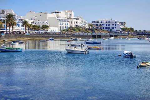 Blue water with boats in the Charco de San Ginés lagoon in front of a coastal town with palm trees