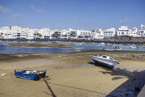 Boats on the banks of Charco de San Ginés lagoon in front of white buildings under blue sky,