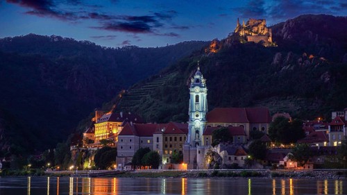 Illuminated church and castle in Dürnstein in front of a hill at dusk, reflected in the river,