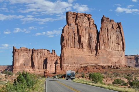 A road leads through a desert landscape with impressive sandstone formations and isolated vehicles