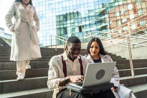 Three diverse business people collaborating outdoors, two using a laptop on city stairs while a