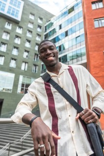 Young black man smiling and leaning on a railing in a modern city street, carrying a bag and