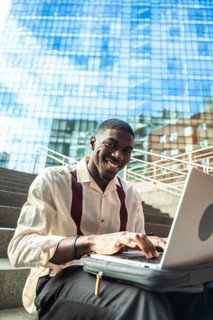 Young black man smiling while typing on a laptop, sitting on urban stairs with a modern glass
