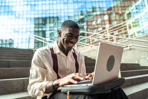 Young black man sitting on concrete stairs in a city, smiling and typing on a laptop, portraying