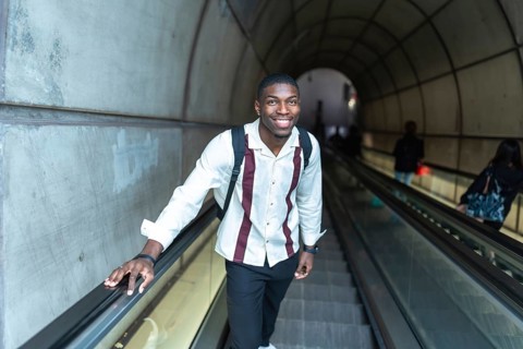 Young black man with backpack smiling at camera while holding handrail on an escalator ascending