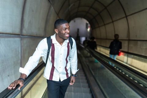 Young black man smiling while traveling up an escalator in a subway station tunnel, carrying a