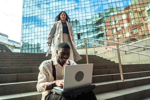 Young professional smiling at laptop on urban steps while a stylish woman walks down nearby by a