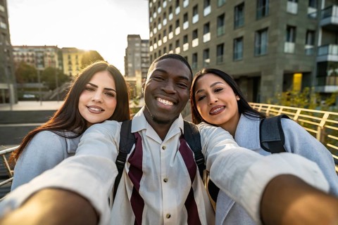 Three diverse students smiling into a smartphone, taking a city street selfie while walking and