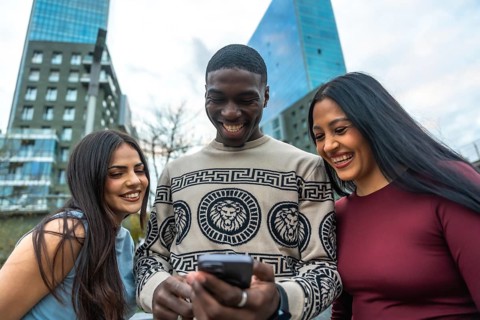 Diverse friends laughing and smiling while gathered on a city street, looking at a smartphone and