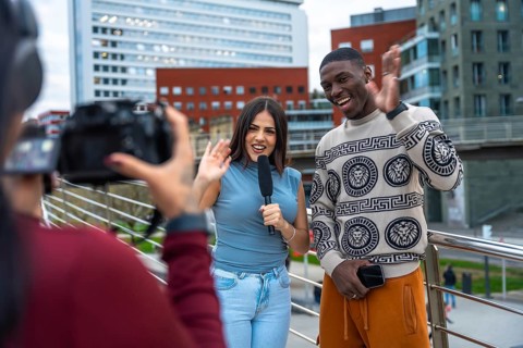 Journalist holding a microphone interviewing a young man on a city bridge, both waving and smiling