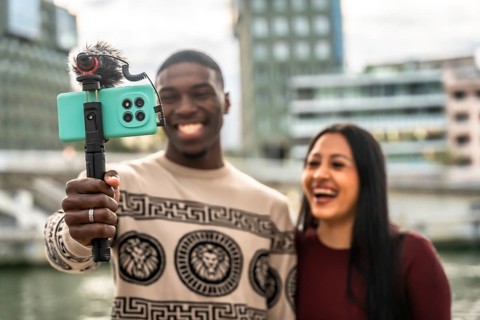 Diverse couple smiling and laughing while creating a vlog, holding a smartphone with an external