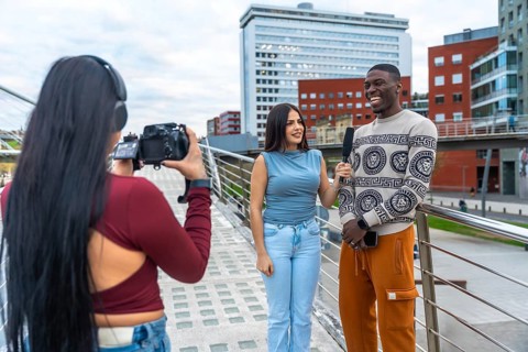 Woman filming an outdoor interview with a young woman holding a microphone to a laughing man,