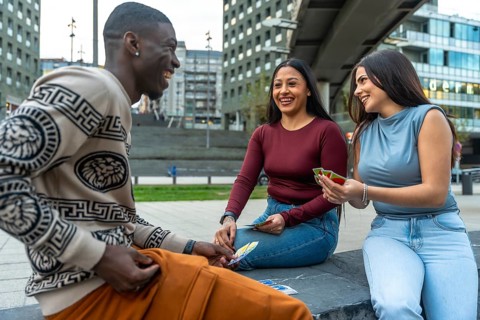 Diverse young friends laughing together while sitting and playing cards outdoors in a modern urban