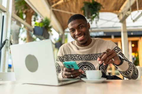 Smiling black man sitting at a cafe with laptop and smartphone, stirring coffee while working