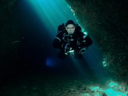 A diver with equipment floats through the interior of a blue-lit cave. Fraskeric dive site, Stoja,