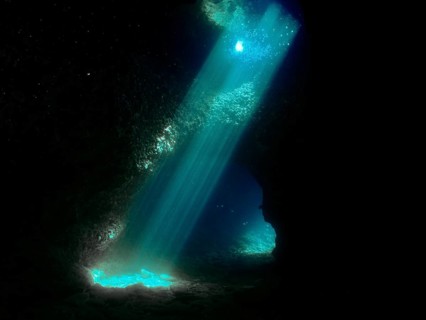 Sun rays illuminate the interior of a dark underwater cave with blue light reflections. Fraskeric