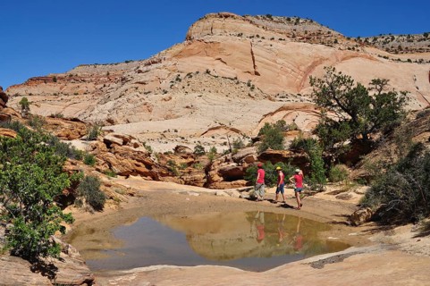 Three hikers explore rocky landscape with small water basin in bright skies, Capitol Reef National