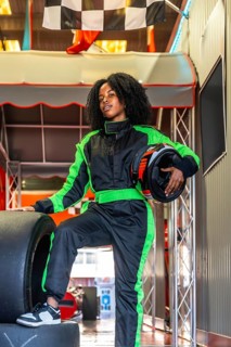 Young black woman standing confidently in a green and black race suit, holding a helmet and resting