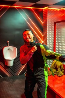Smiling young man in neon lit restroom pulling on black and green karting jumpsuit, getting dressed